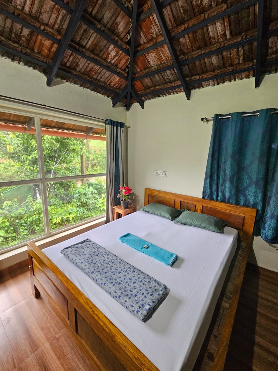 Teak double bed under a tiled roof with a large window facing the green plantation